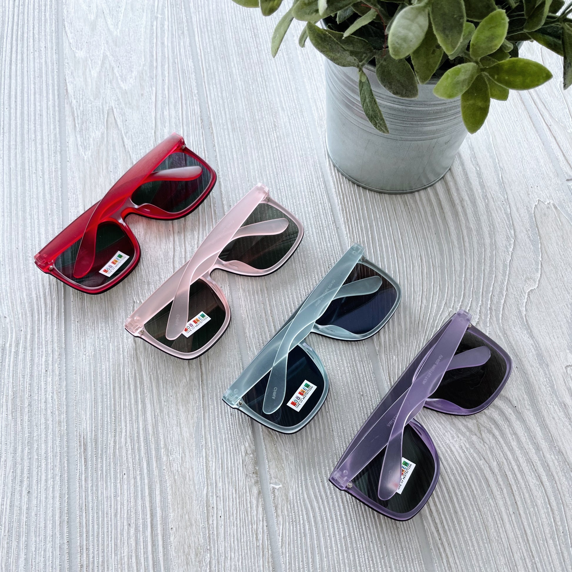 Four pairs of Arden Mirrored Sunnies • Kids in red, pink, blue, and purple lie crossed on a light wooden surface next to a potted green plant. Each pair features a small lens label and is available as final sale.