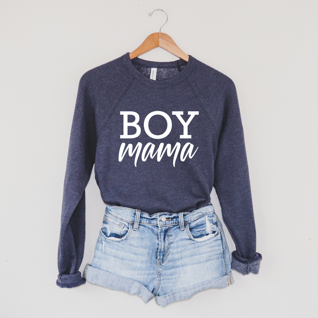 The Boy Mama • Navy Pullover hangs on a wooden hanger above light blue denim shorts, displayed against a plain light-colored wall.