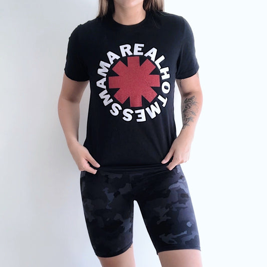 A person stands against a plain background wearing a black T-shirt that says MAMA REAL HOT MESS with a red asterisk, paired with Charcoal Camo • Brushed Biker Shorts. Their hands rest on their hips.