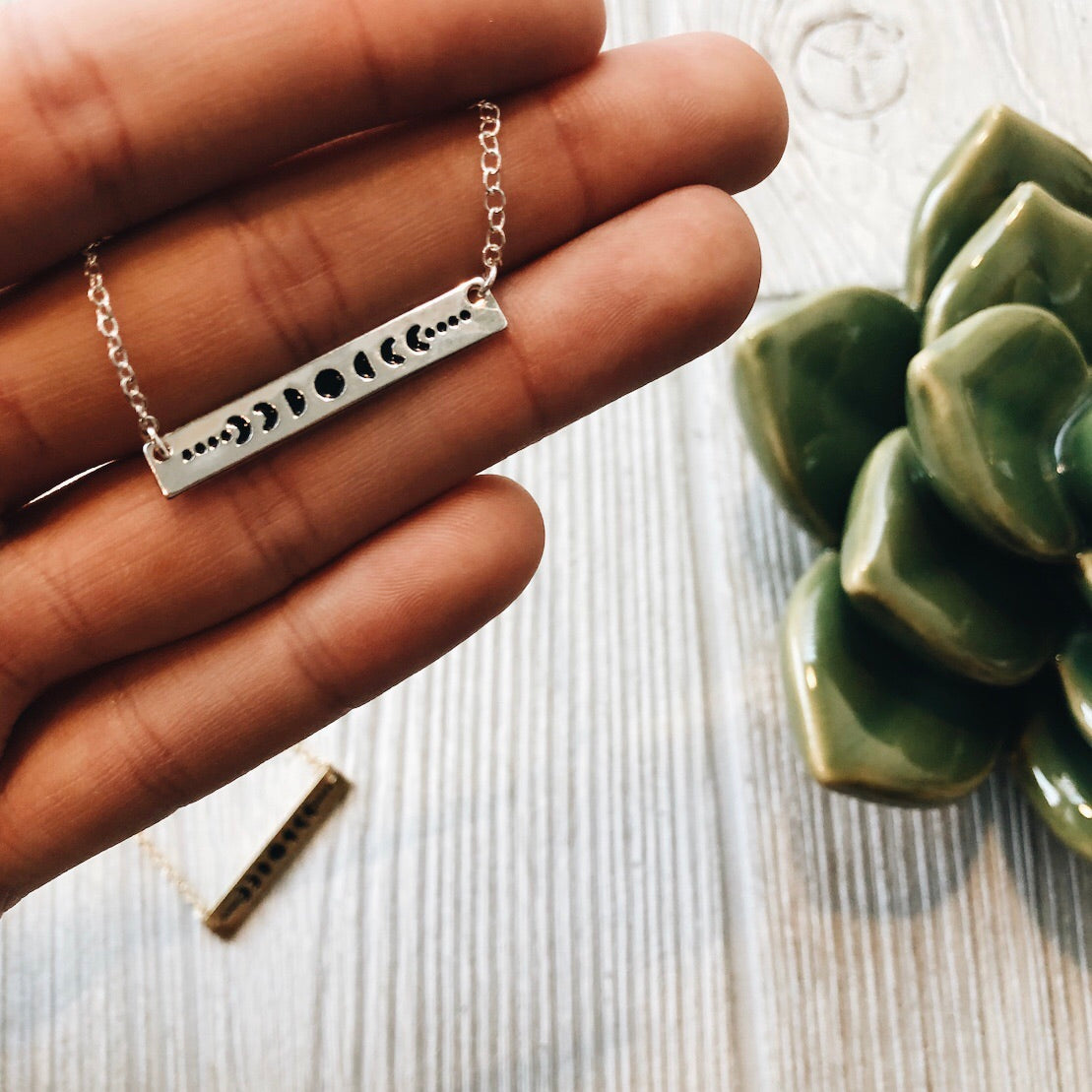 A hand holds the Moon Phase • Bar Necklace, featuring an adjustable clasp. In the background, a green succulent and another silver or gold necklace lie on a light wooden surface.