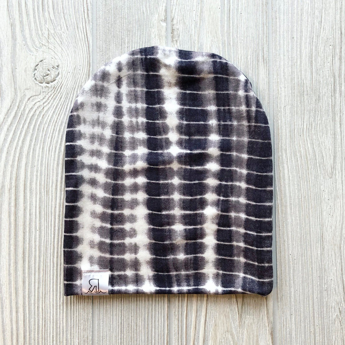 The Black Dyed Beanie, featuring a black and white grid tie-dye pattern and soft interlock knit, is laid flat on a light wooden surface with a small white tag displaying a black R logo on the bottom left edge.