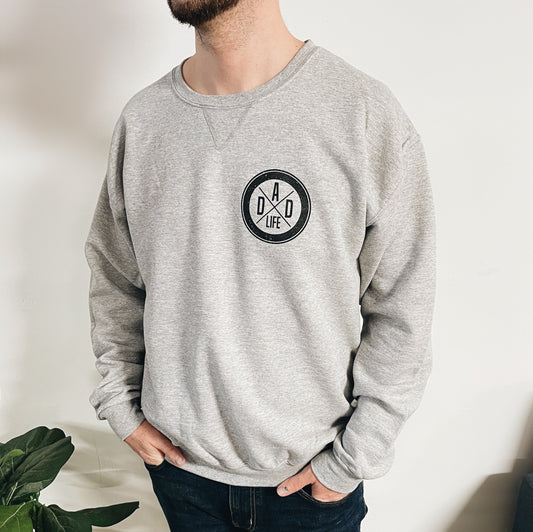 A man stands with his hands in his pockets against a white wall, wearing the Dad Life • V-Notch Pullover in light gray, featuring a round Dad Life logo on the chest. A green plant appears partially at the bottom left.