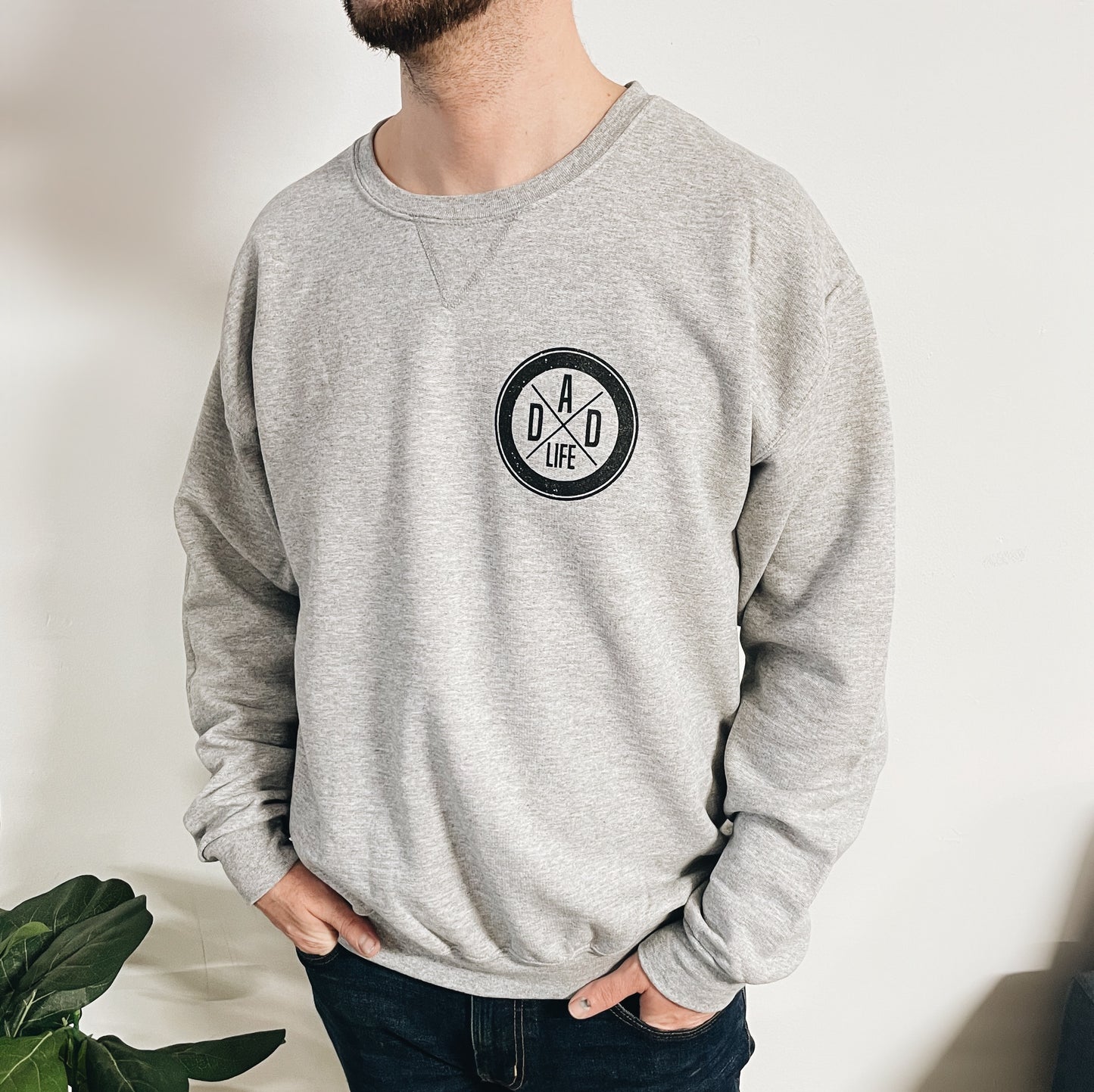 A man stands with his hands in his pockets against a white wall, wearing the Dad Life • V-Notch Pullover in light gray, featuring a round Dad Life logo on the chest. A green plant appears partially at the bottom left.