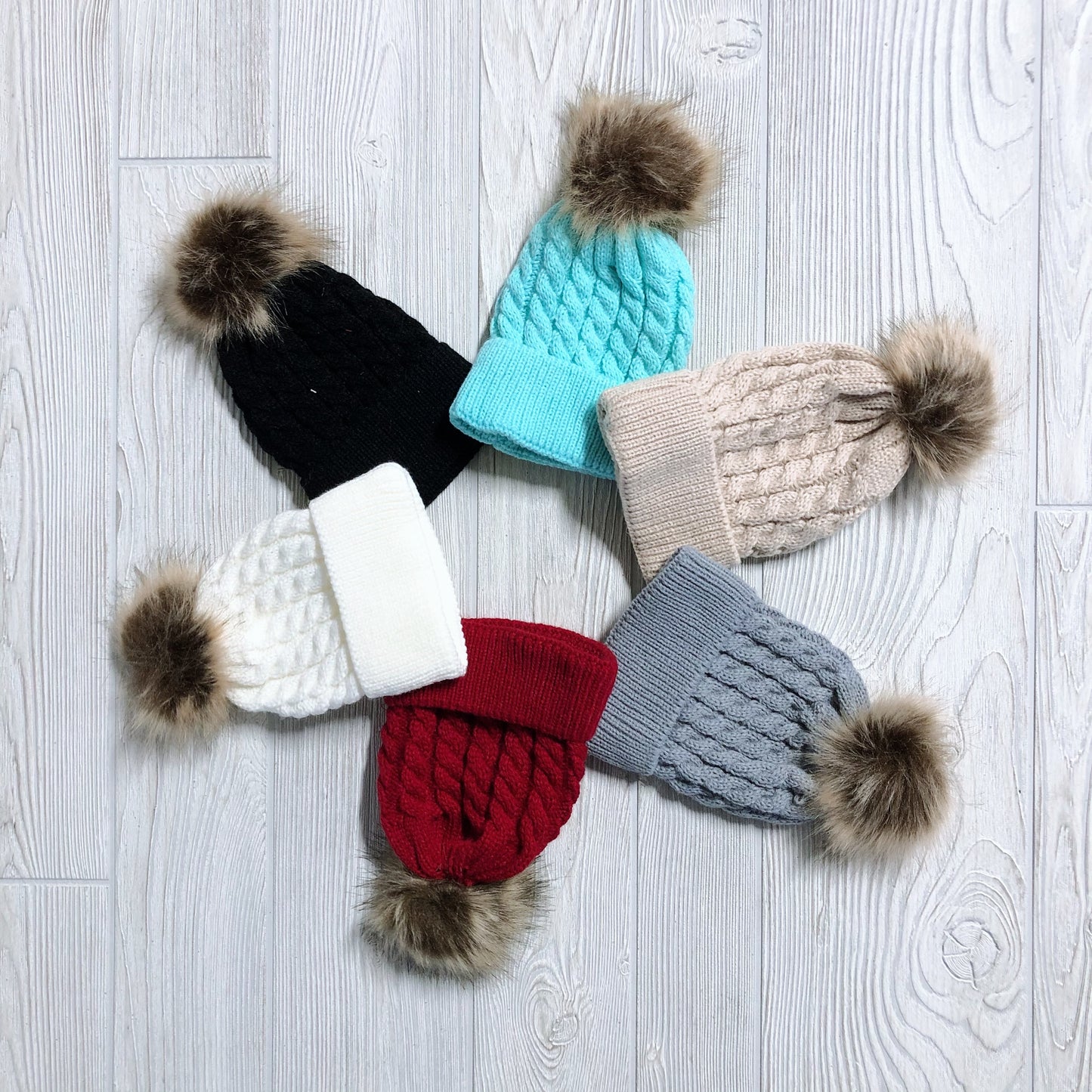 Six Cable Knit Pom Beanie • Infant hats, in black, light blue, beige, gray, red, and white, are arranged in a circle on a light wooden floor. These warm knitted hats with fur pom-poms are perfect cold weather accessories for infants.