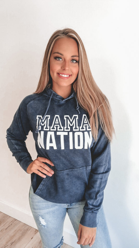 A woman with long blonde hair smiles at the camera, wearing a dark blue Mama Nation • Vintage Hoodie and light ripped jeans, standing against a plain white background.