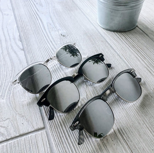 Three pairs of Zoe Mirror Sunglasses in assorted colors, featuring reflective lenses, are displayed on a light wood surface beside a silver pot. Plant reflections are visible in the lenses. Final Sale—Not Eligible for Return.