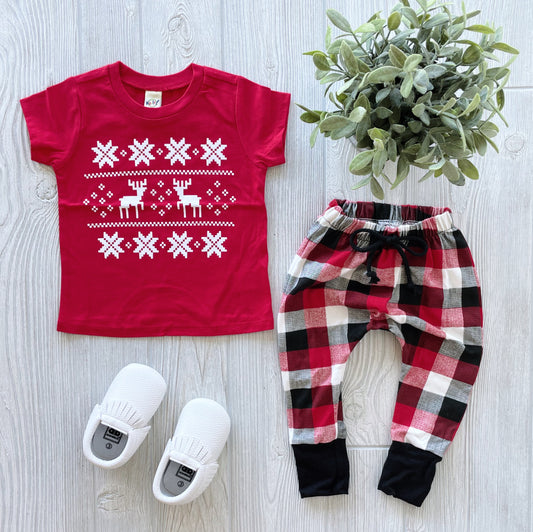 A red Ugly Sweater • Kids Tee with white reindeer and snowflakes, paired with red and black plaid pants, white baby shoes, and a green potted plant displayed on a light wood surface.