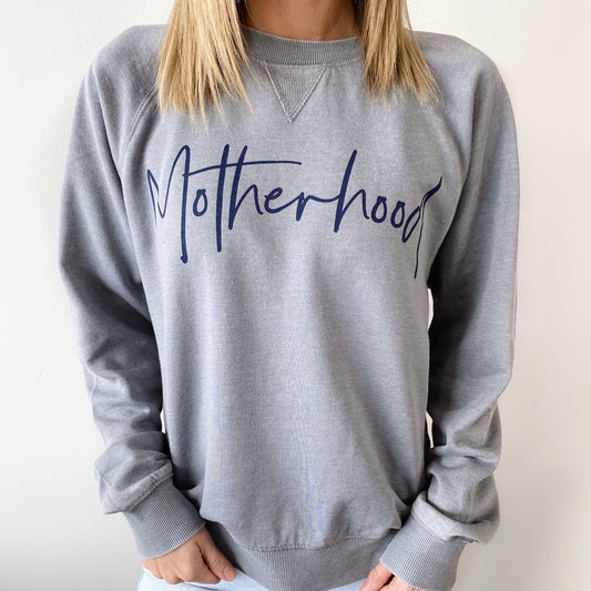 A woman wears the Motherhood Script • Gray Vintage Fleece sweatshirt, featuring Motherhood in bold navy cursive across the chest. The light gray fleece has a relaxed unisex fit, and she has straight, shoulder-length hair.