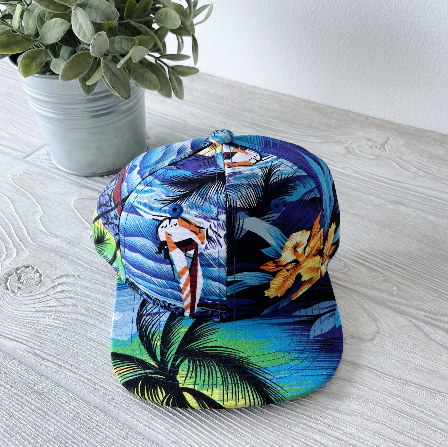 A Kids Snapback • Wyatt featuring beach, palm tree, and flower patterns rests on a light wood surface beside a potted green plant.