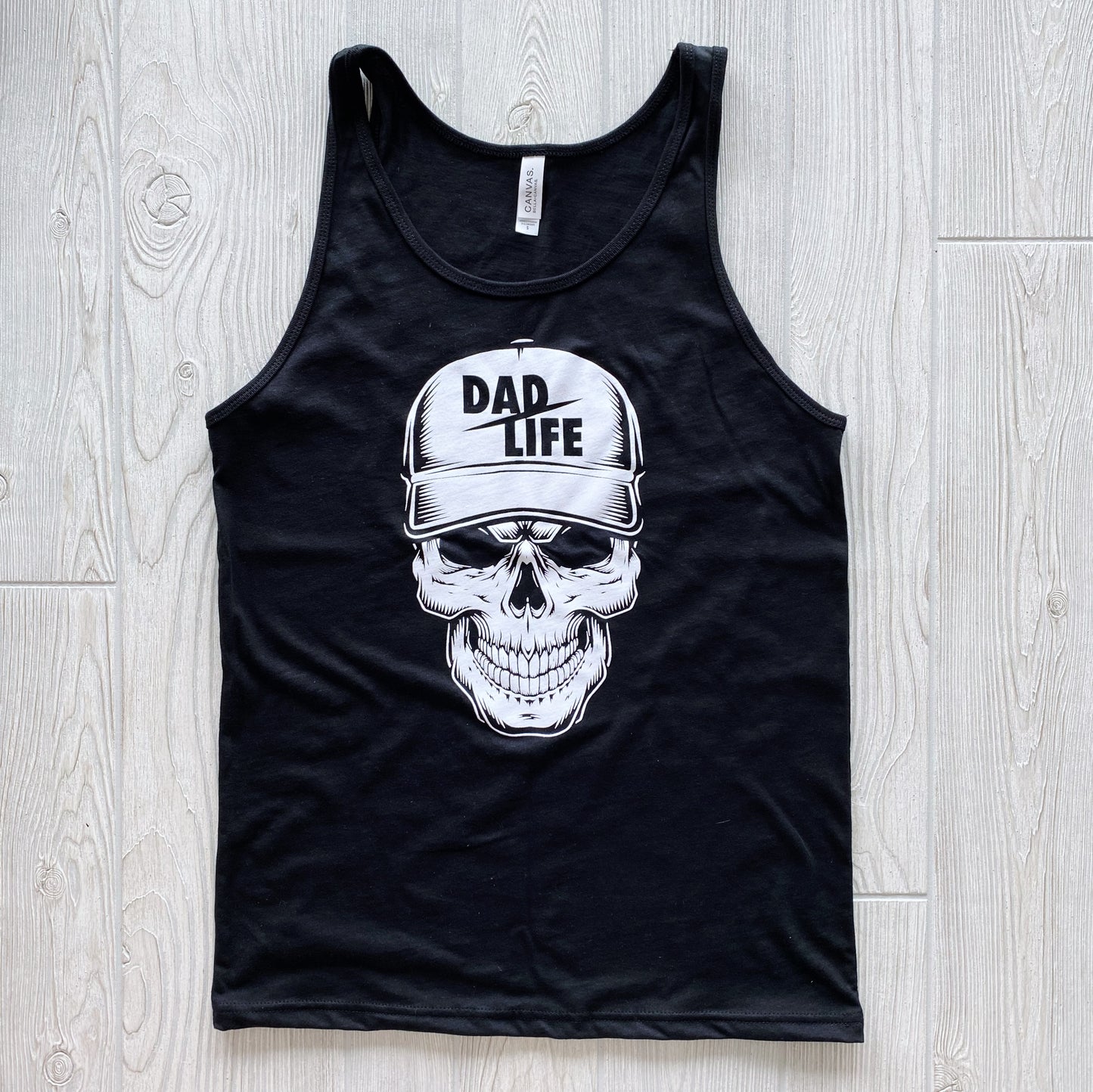 The Skeleton Dad • Black Tank is a Bella/Canvas unisex tank top in black, featuring a bold skull with a “DAD LIFE” baseball cap, displayed flat on a light wood floor.