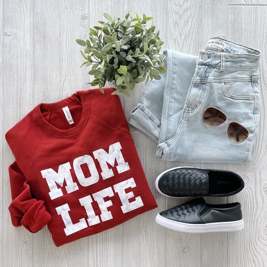 Flat lay of MOM Marble • Brick Pullover—a red sweatshirt with bold white MOM LIFE letters—paired with light-wash jeans, black slip-ons, brown sunglasses, and a potted plant on a light wood floor. Relaxed unisex fit.