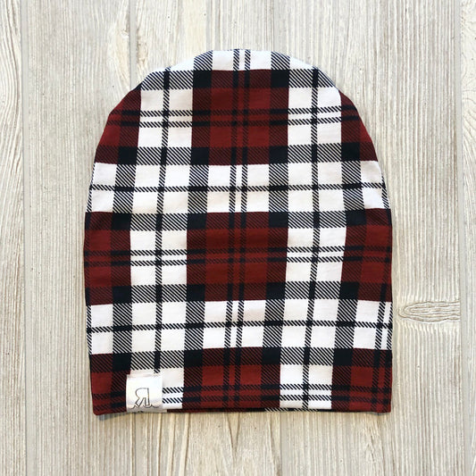 A Burnt Red Plaid Beanie is laid flat on a light-colored wooden surface, highlighting its cozy plaid knit pattern in shades of red, black, and white.