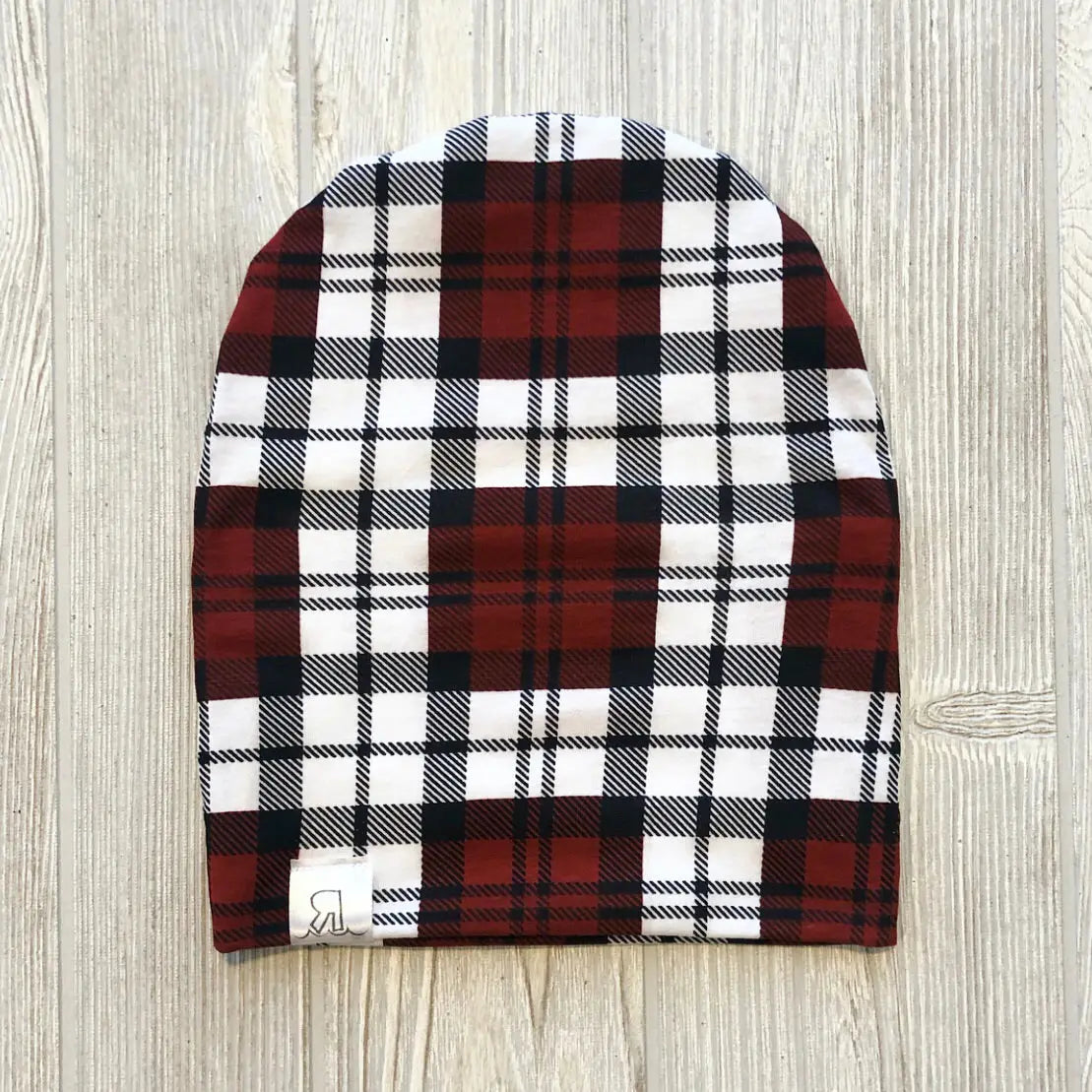 A Burnt Red Plaid Beanie is laid flat on a light-colored wooden surface, highlighting its cozy plaid knit pattern in shades of red, black, and white.