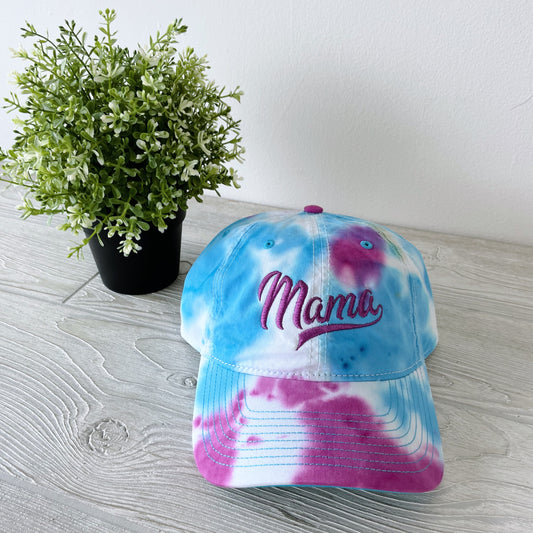 The Tie Dye Hat • Jelly Bean, a vintage-style unisex cap with Mama embroidered in pink, rests on a light wooden surface beside a small potted plant, showcasing its colorful tie-dyed twill.
