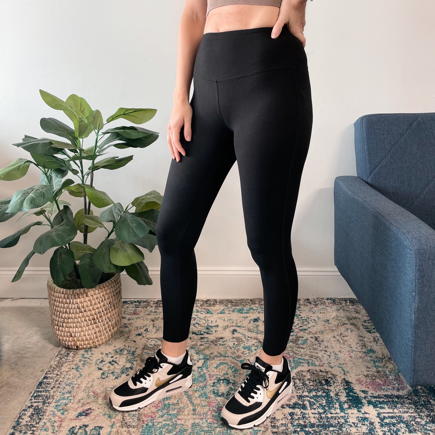 Wearing Lydia Leggings and black-and-white sneakers, a person stands indoors near a green potted plant and blue couch on a patterned rug, with only the lower half of their body visible.