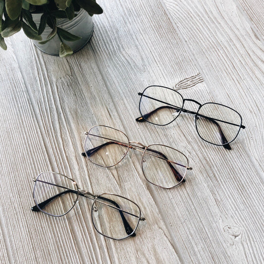 Three pairs of Frankie • Blue Light Glasses in silver, gold, and black are displayed on a light wooden surface with a potted green plant nearby—ideal for reducing headaches and eye strain. More colors available!.
