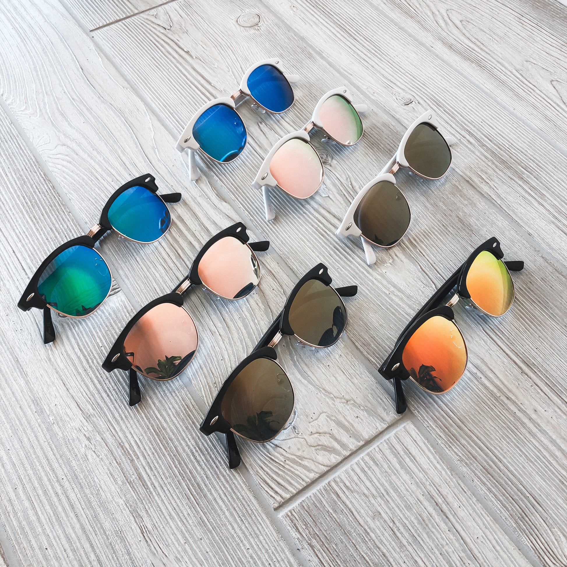 Seven pairs of Mirror Wayfarer Sunglasses (Adult) with multicolored reflective lenses—blue, pink, green, orange, and brown—are arranged on a light wooden surface. Each stylish pair offers UV400 protection for sun safety. Available in more colors!.