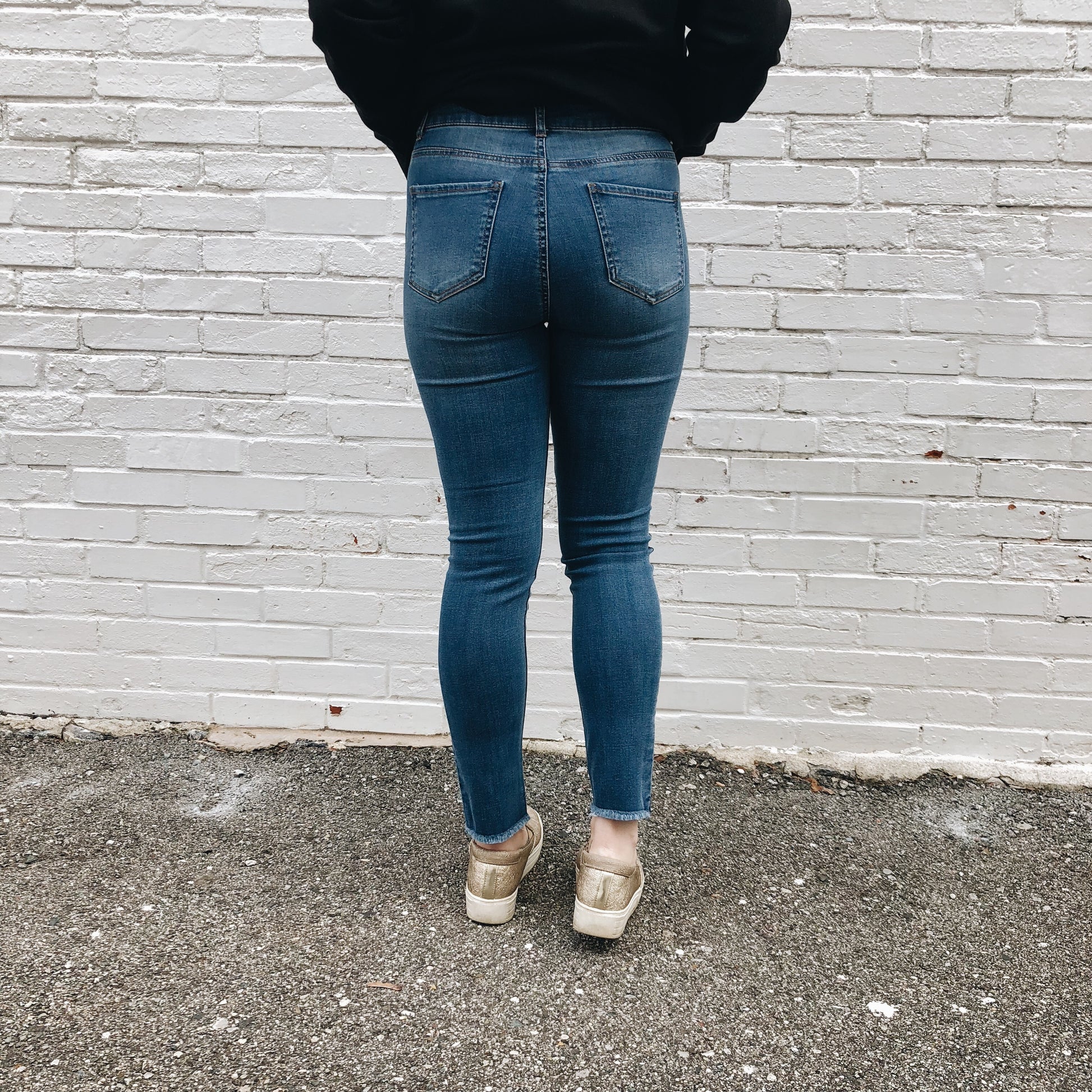 A person stands facing a white brick wall, shown from behind (shoulders to feet), wearing Alie Jeggings SIZE 0 & 3, a black top, and beige shoes on asphalt. The jeggings are skinny fit with super stretch and true-to-size.