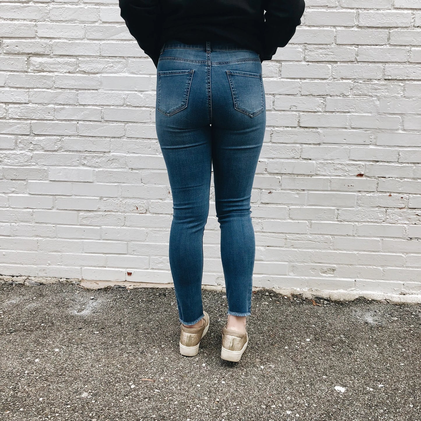 A person stands facing a white brick wall, shown from behind (shoulders to feet), wearing Alie Jeggings SIZE 0 & 3, a black top, and beige shoes on asphalt. The jeggings are skinny fit with super stretch and true-to-size.