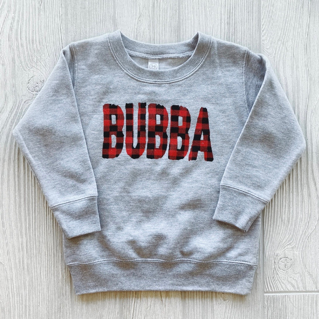 The Bubba Plaid • Pullover is a gray long-sleeve sweatshirt featuring BUBBA in bold red and black buffalo plaid letters on the front, shown on a light wood background.