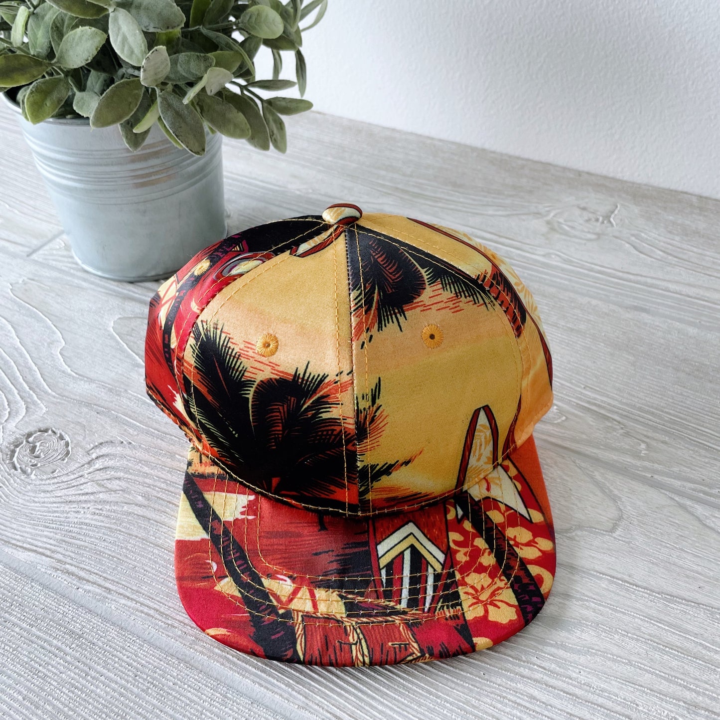 A Kids Snapback • Levi with a tropical design of palm trees and surfboards rests on a light wooden surface beside a small potted plant.