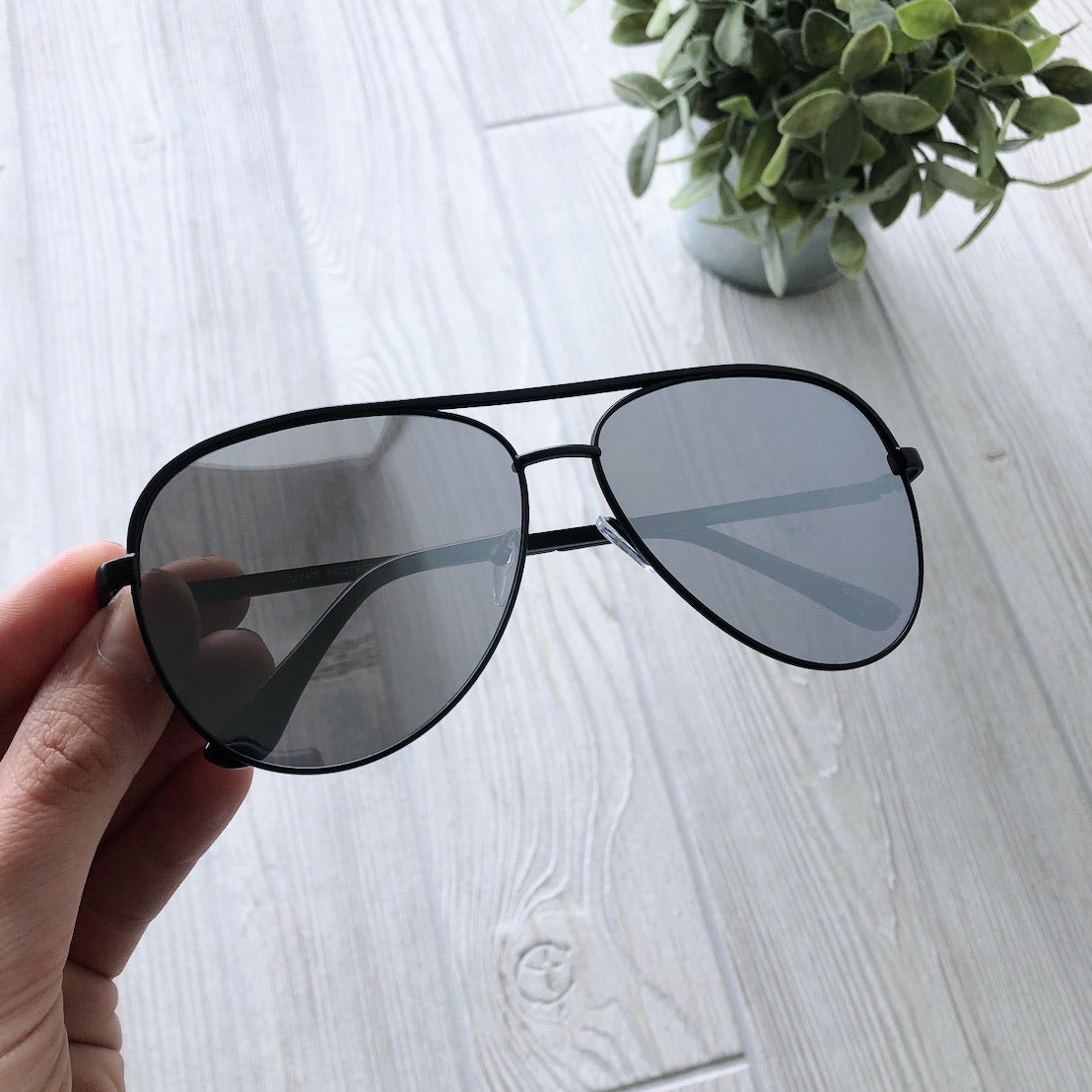 A hand holds Avery Sunglasses • Adult, featuring black aviator frames and UV400 dark lenses, above a light wood floor with a potted green plant in the background.