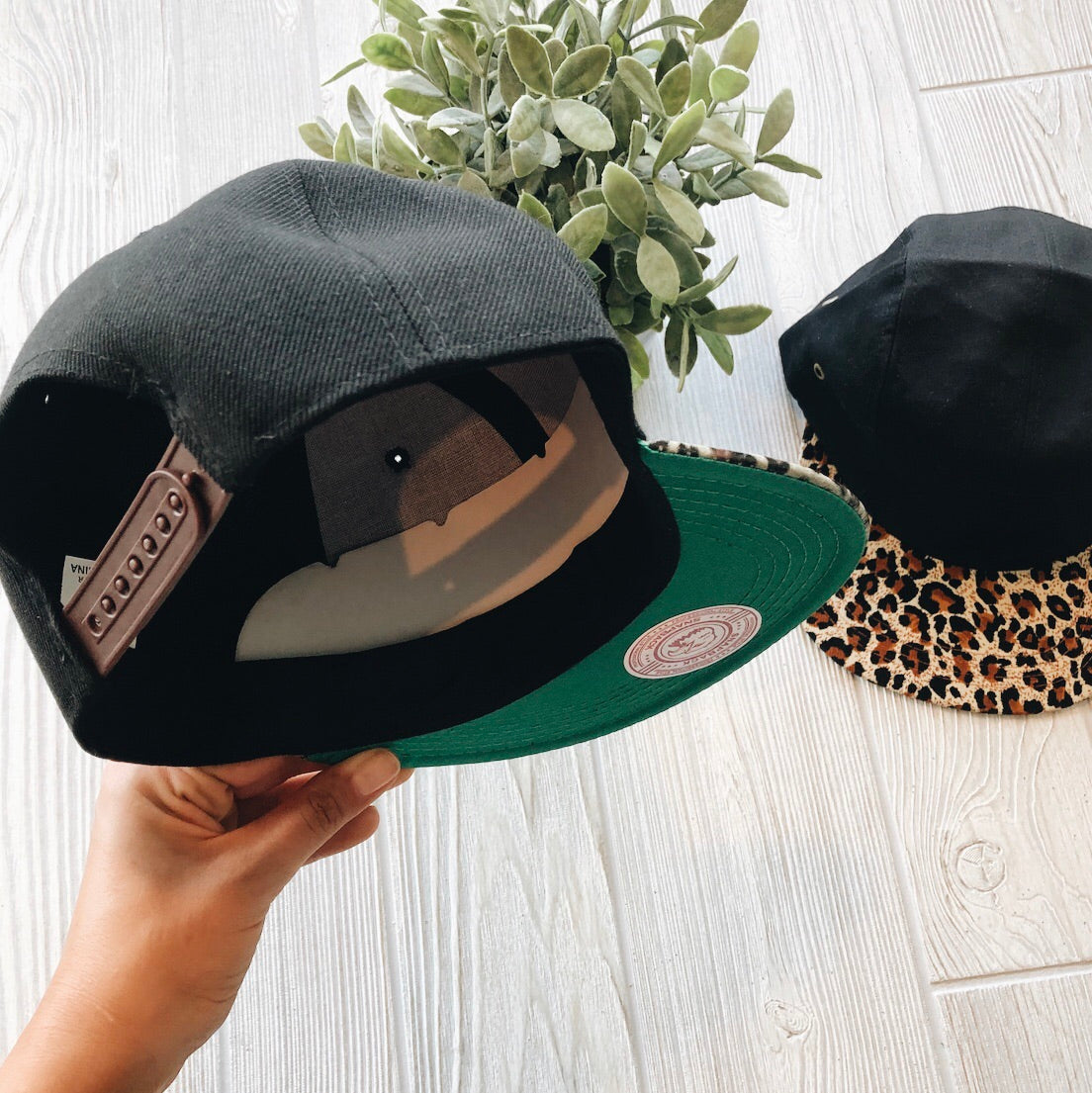 A hand holds a black snapback with a green brim underside and adjustable strap. On the table are a green plant and the Mommy & Me • Cheetah Hat, featuring a leopard print brim, set against light wood in the background.
