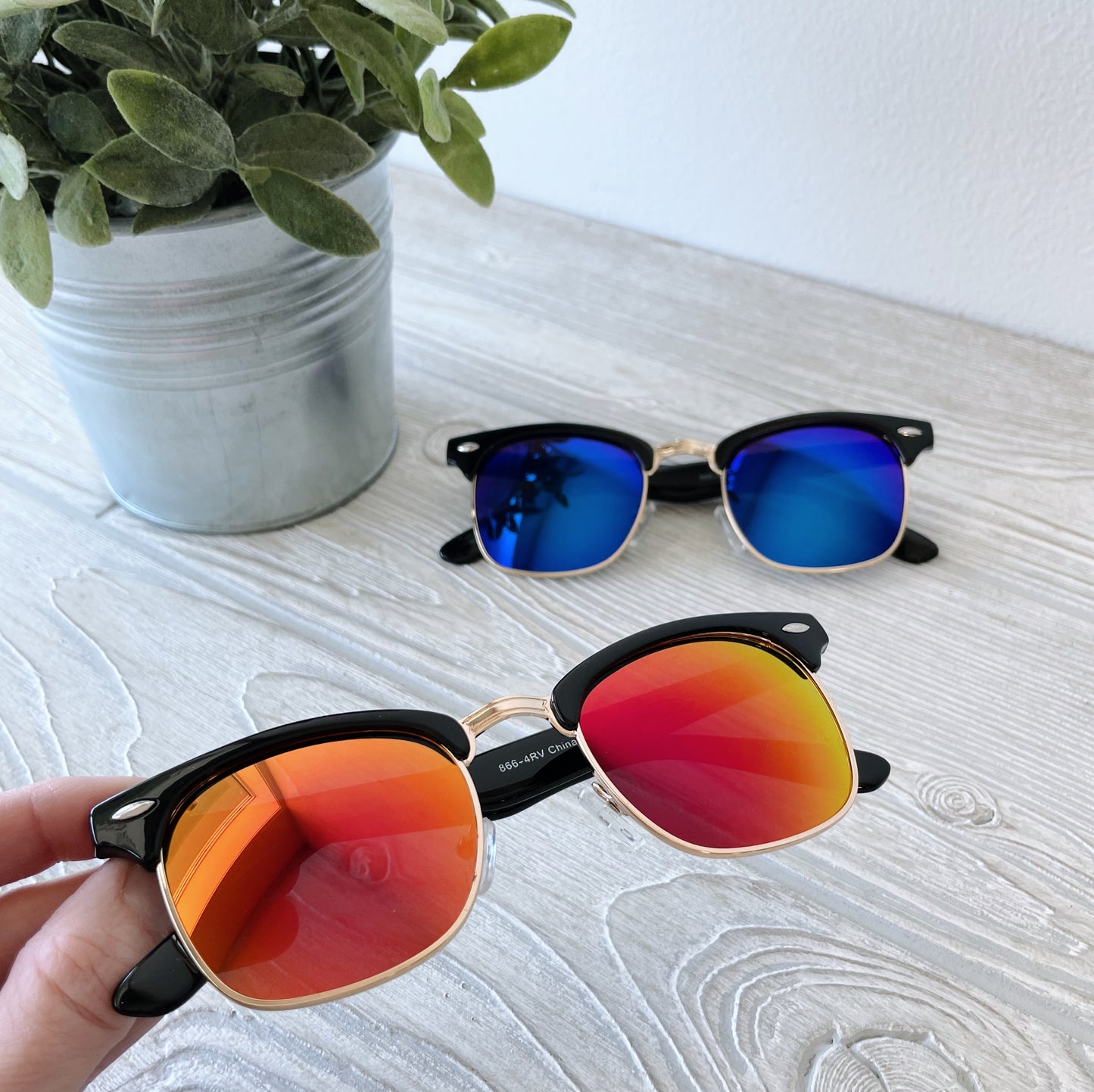 A hand holds Parker • Adult Sunglasses with red-orange mirrored lenses above a light wood surface; in the background, blue-lensed sunglasses and a potted plant appear. Final Sale – don’t miss out!.