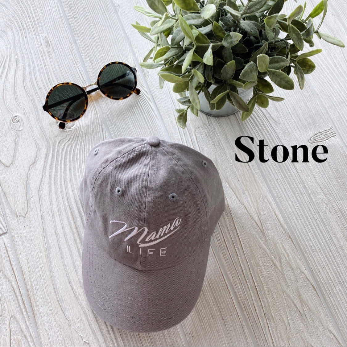 A gray Mama Life Hat • NEW Colors! with vintage white embroidery rests on a light wood surface beside round sunglasses and a potted green plant. Stone is displayed on the right side of the image.