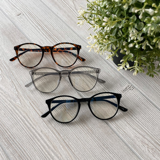 Three pairs of Temp Blue Light Glasses in tortoiseshell, clear, and black rest on a light wood surface beside a leafy potted plant, offering stylish protection from eye strain. More colors available!.