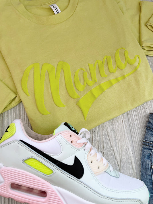 The Sporty Mama • Neon Tonal Long Sleeve, a pale yellow tee with Mama in large script and sporty style, is displayed beside a white Nike sneaker featuring pastel pink, yellow, and black accents on a light wood surface.