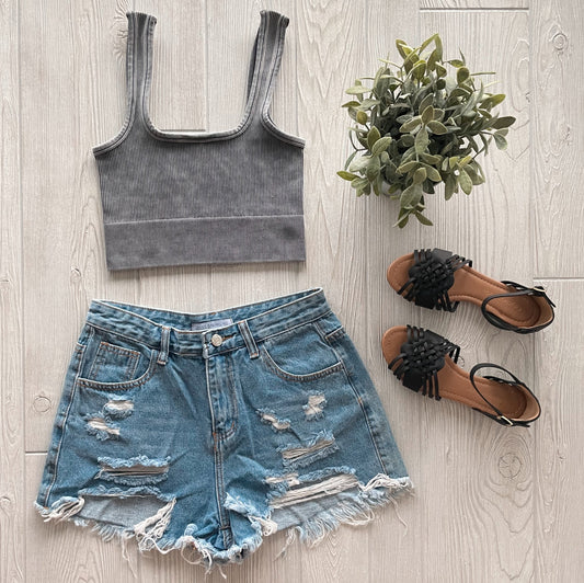 Flat lay of a summer outfit featuring the Iris Crop from the Denver Collection, paired with distressed denim shorts, black braided sandals, and a small green potted plant on a light wooden floor.