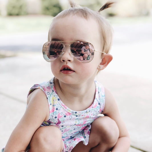A young child with short hair and pigtails wears Aviator Sunnies • Kids and a colorful sleeveless dress, crouching outdoors on a sunny day. The reflective sunglasses show the photographer’s image.