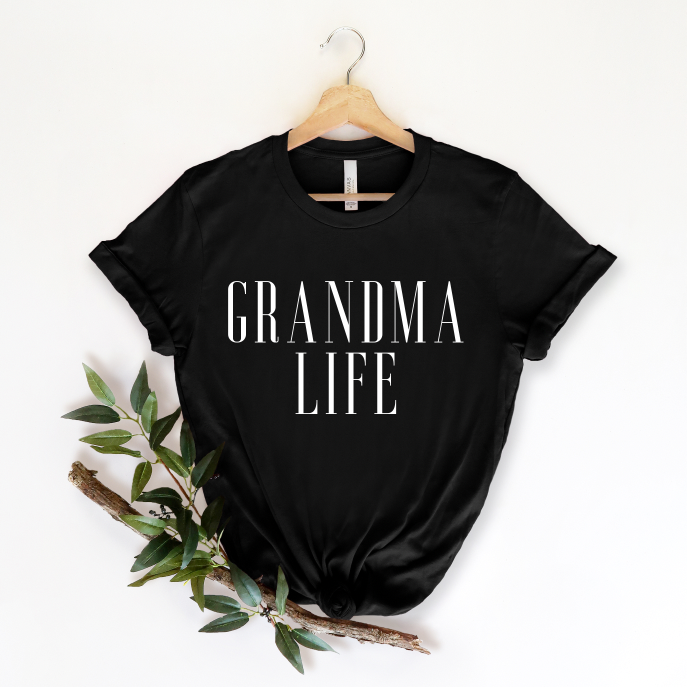 The GRANDMA LIFE • Black Tee is a unisex black shirt featuring GRANDMA LIFE in bold white lettering, displayed on a hanger with a leafy branch underneath.