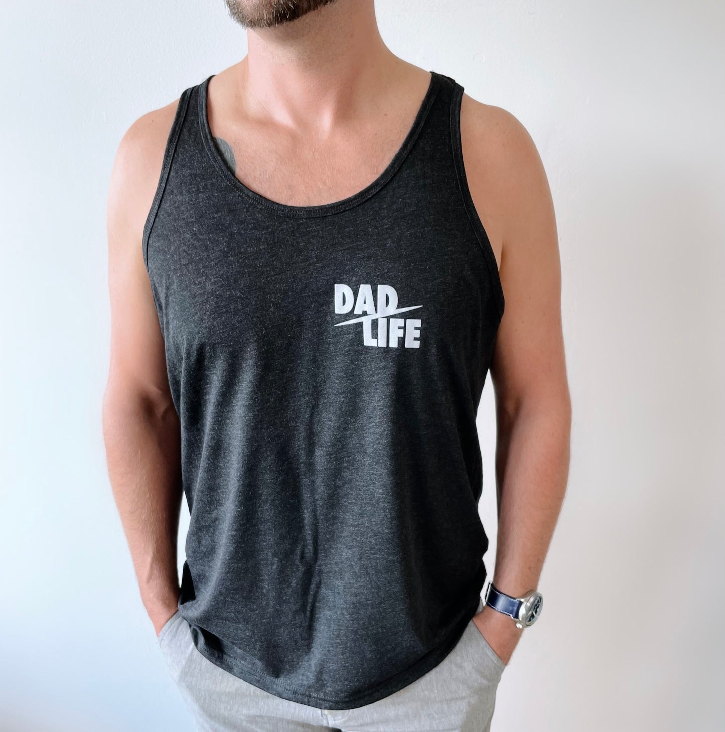 A man stands against a white background with his hands in his pockets, wearing the Dad Life • Tank—a black Bella/Canvas unisex tank top featuring “DAD LIFE” printed in white on the chest.