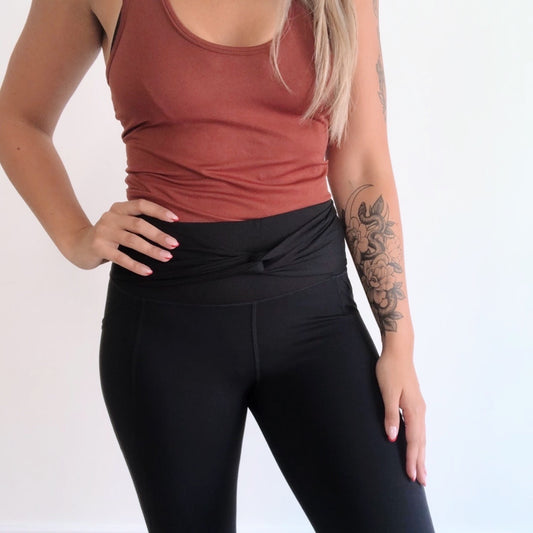 Wearing a sleeveless rust top and Twist Accent Highwaist Legging • 2869 in black, a person stands against a white background with one hand on their hip, showing a floral tattoo on their left forearm.