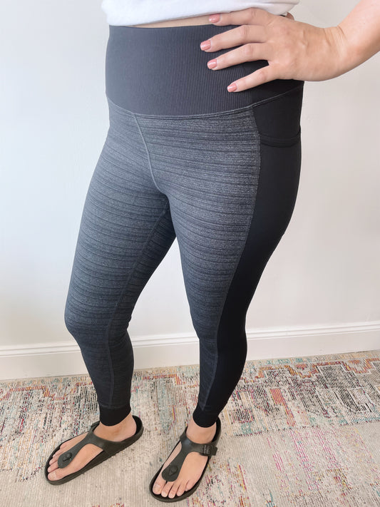 Person wears Cuffed Leggings • 6191 in gray and black with tummy control, a white top, and dark green sandals while standing on a multicolored textured rug, one hand resting on their hip.