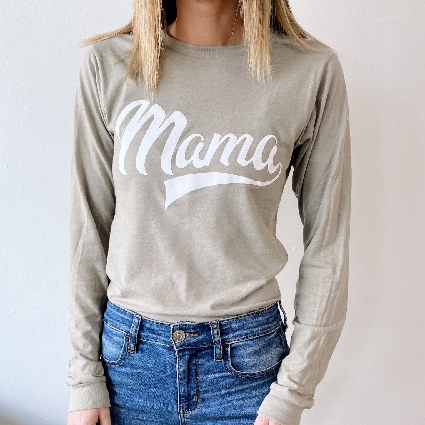 A woman wears the Mama Sporty • Stone Long Sleeve, a beige top with a bold white Mama print across the chest, paired with blue jeans. Her face is not visible.