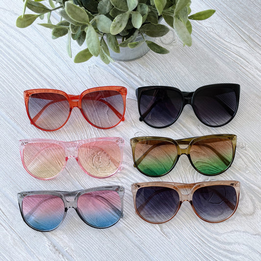 Six pairs of True • Adult Sunglasses with large, round frames in assorted shades and gradients are displayed in two rows on a light wood surface, topped by a potted green plant.