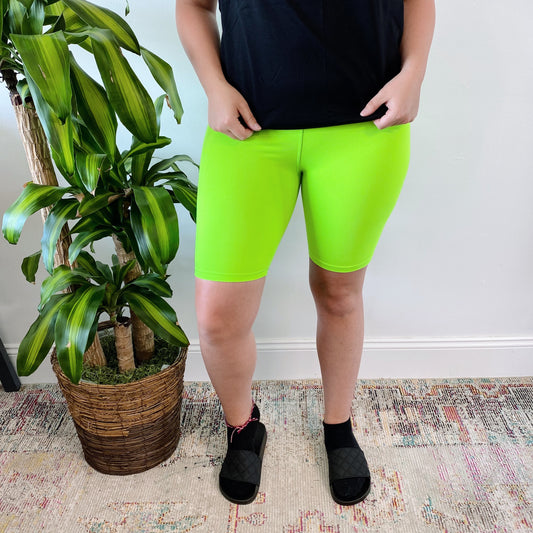 A person stands indoors on a patterned rug next to a leafy potted plant, wearing Lime • Brushed Biker Shorts, a black shirt, and black slide sandals. Their head is not visible in the image.