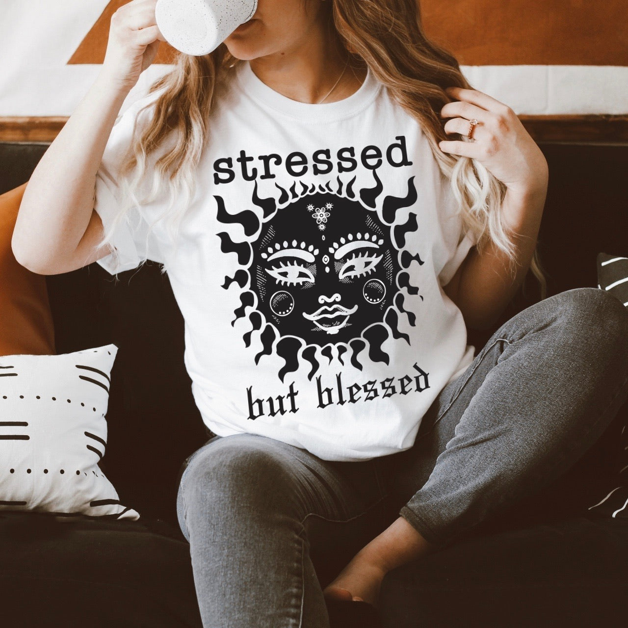 A woman with long, wavy hair sits on a couch, sipping from a white mug. She wears the Stressed • White Tee, featuring an illustrated sun face and unisex fit. Geometric patterned pillows are beside her.
