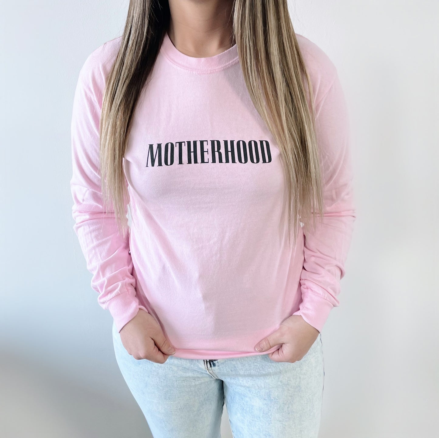 A woman wears the Motherhood • Pastel Pink Long Sleeve, a soft ring-spun cotton shirt featuring bold black MOTHERHOOD text on the front, standing against a plain background with her hands in her jean pockets.