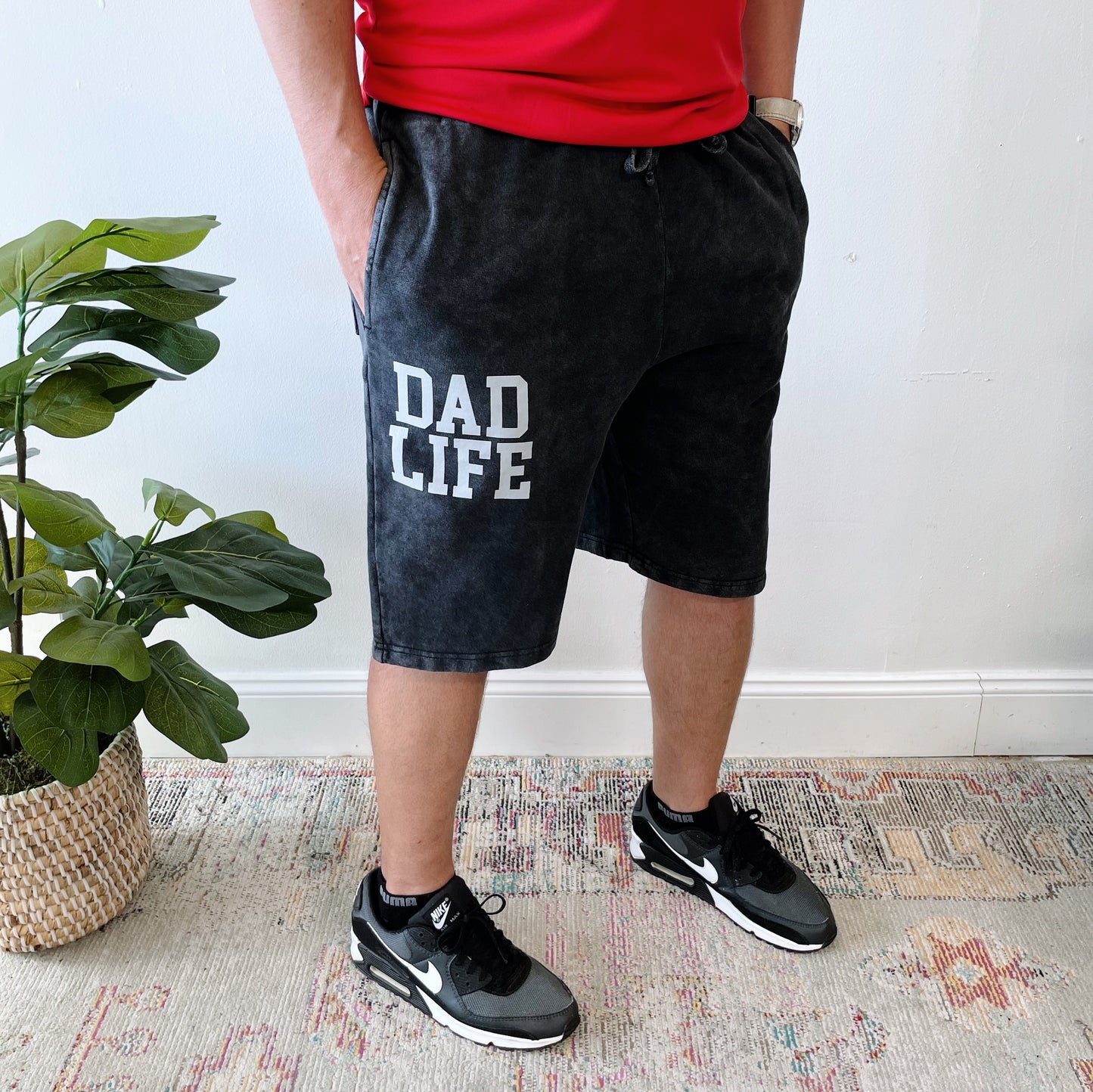 A man wears Dad Life Shorts • Acid Wash, featuring a drawstring waist and DAD LIFE on the left leg, paired with a red shirt and black Nike sneakers. He stands indoors by a potted plant and rug for a relaxed loungewear vibe.