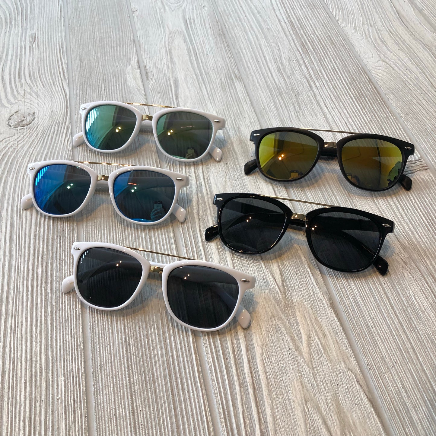 Five pairs of Wayfarer Bar Sunglasses (Adult Size, More Colors!) with UV400 protection are arranged on a light wooden surface; three have white frames, two have black frames, and the lenses and frames come in different colors.