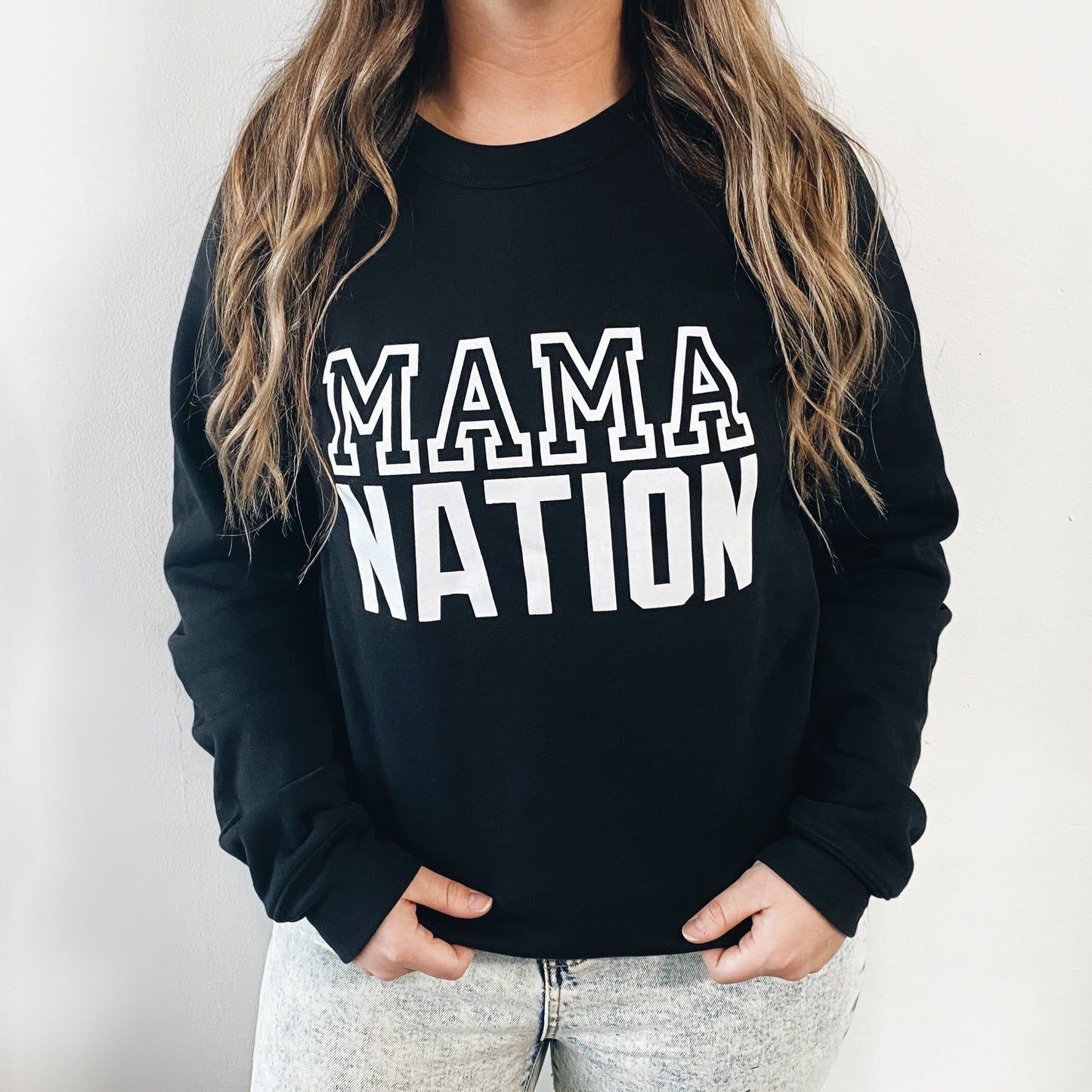Someone wears the MAMA NATION • Black Pullover, a unisex sweatshirt featuring bold white lettering. Their long wavy hair complements the look as they stand against a plain white background.