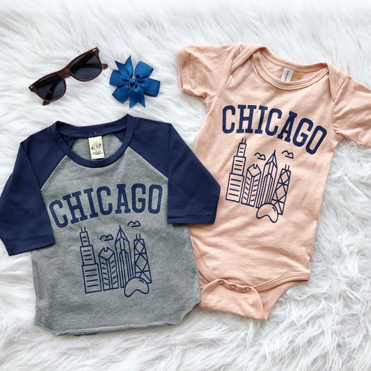 Two Chicago • Raglan/Bodysuit baby outfits—one gray-blue raglan, one peach onesie with city skyline graphics—are displayed on a white fluffy surface beside brown sunglasses and a blue bow.