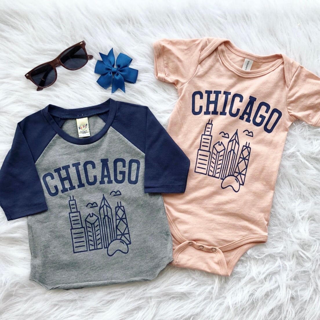 Two Chicago • Raglan/Bodysuit baby outfits—one gray-blue raglan, one peach onesie with city skyline graphics—are displayed on a white fluffy surface beside brown sunglasses and a blue bow.