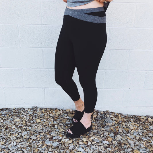 A person stands against a white brick wall, wearing Wrap Band Leggings • 1070 with a unique waistband design and black sandals. Their legs are crossed at the ankles over wood chips.