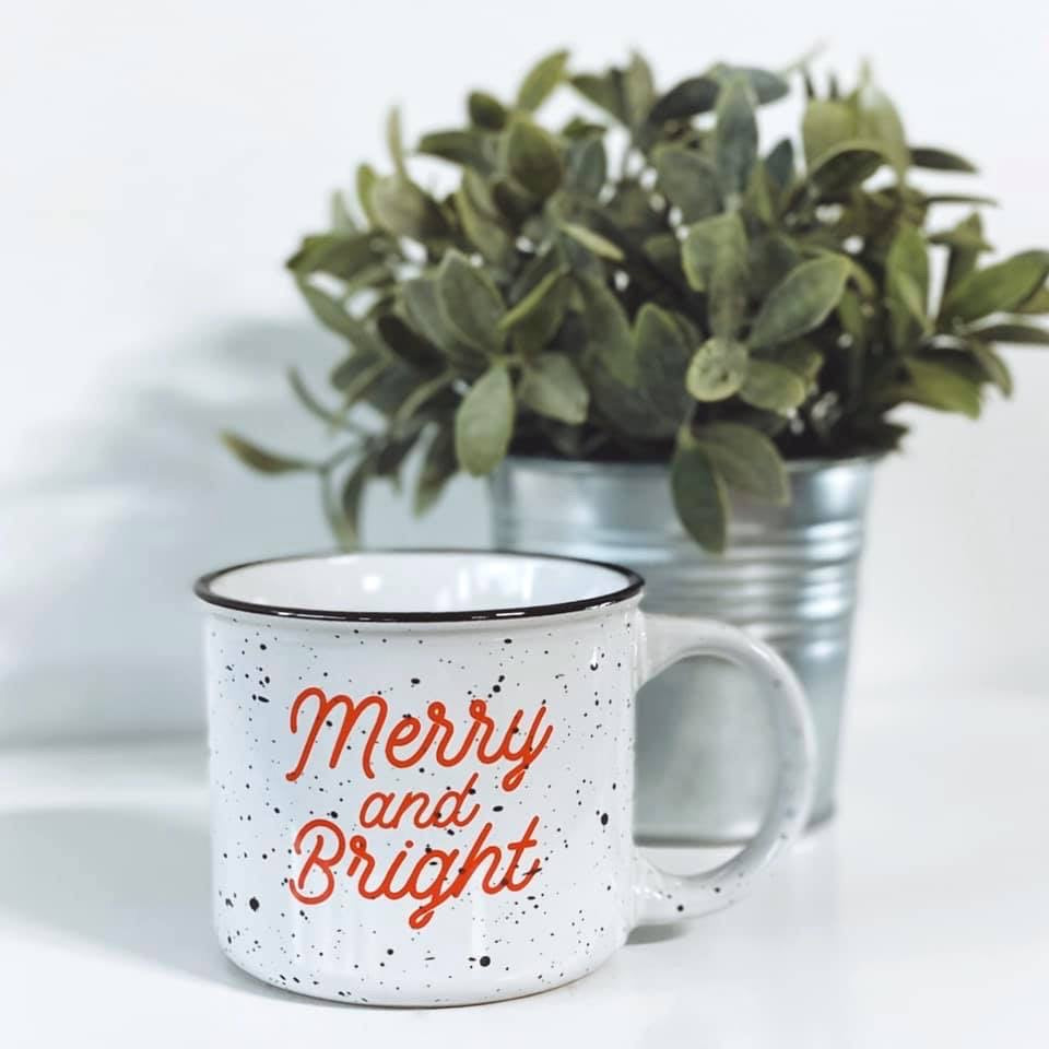 The Merry & Bright • Campfire Ceramic Mug features a white speckled ceramic design with red lettering, displayed in front of a potted green plant in a metal container on a white surface. Hand-wash only.
