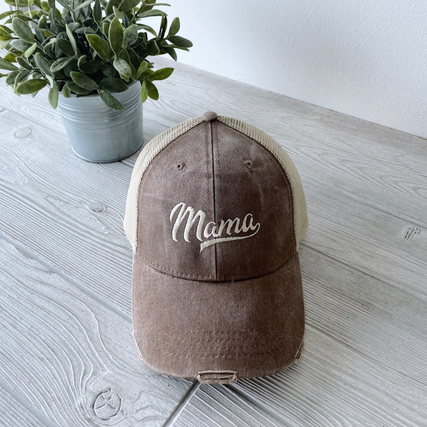 A Distressed Mama Hat in Mocha, featuring white cursive embroidery, rests on a light wood table beside a small potted plant, highlighting the charm of stylish embroidered hats.